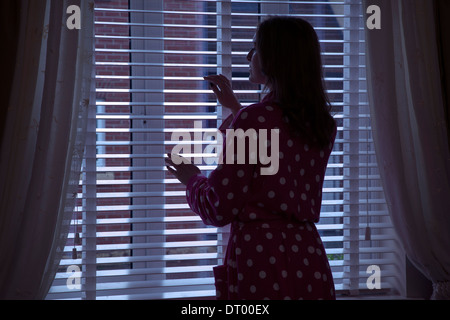 Silhouette of woman wearing a dressing gown looking out of a window at night. Over shoulder back/side view. Stock Photo