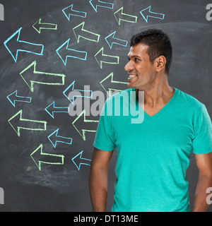 Happy Indian man standing in front of a dark chalkboard with arrow signs drawn pointing. Stock Photo