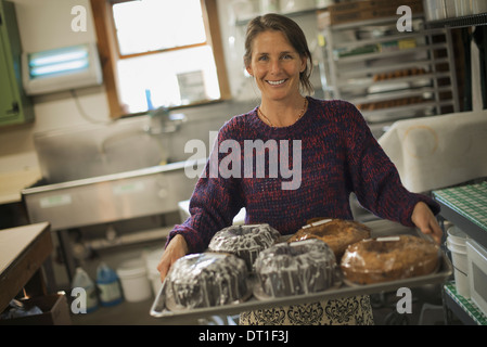 A woman in a kitchen carrying a tray of iced fresh baked cakes Stock Photo