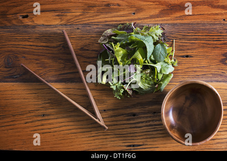 A small round polished wooden bowl and a clutch of organic mixed salad leaves with wooden chopsticks Stock Photo