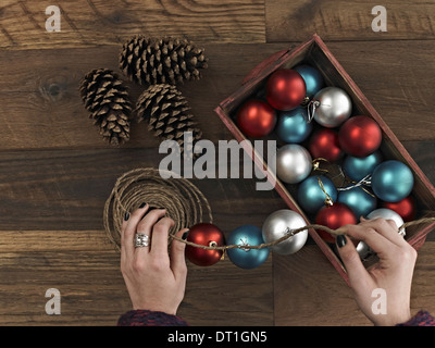 A woman threading Christmas shiny round ornaments on a piece of string A small group of pine cones Stock Photo