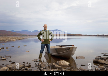 A man standing on a lake shore beside a small rowing boat Stock Photo