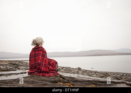 A woman looking out over the water on the shores of a calm lake wrapped in a tartan rug Stock Photo
