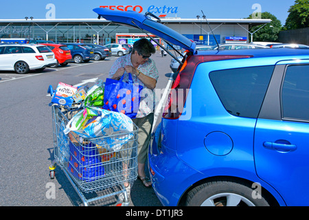 Tesco Extra supermarket store free car park mature woman load food shopping baskets from trolley into open hatchback car tail gate London England UK Stock Photo