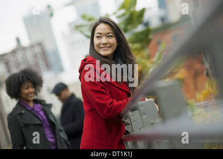 A woman in a red winter coat leaning on a railing Two people in the background City landscape of buildings