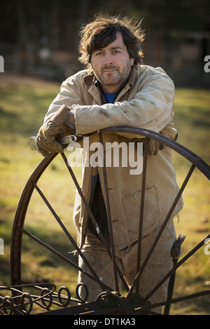 Working on an organic farm A man in overalls wearing work gloves leaning on a round metal wheel with spokes Stock Photo