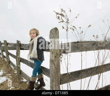 A farm in the snow A girl with a hat and scarf and boots leaning on a fence Stock Photo