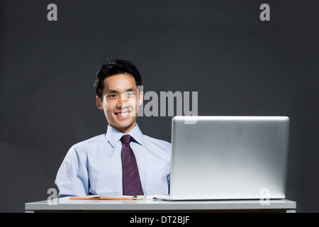 Portrait of an Asian business man sitting at a desk working with a laptop. Dark grey background Stock Photo