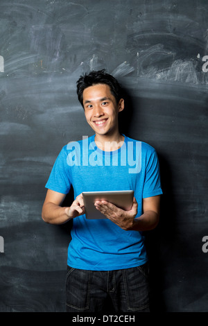Thoughtful Asian man using a tablet PC standing next to a blackboard. Stock Photo