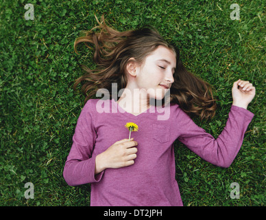 A ten year old girl lying on the grass with her eyes closed holding a dandelion flower Stock Photo