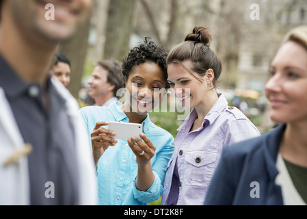 People outdoors in the city spring time New York City park Two women in a group of friends looking at a cell phone and smiling Stock Photo