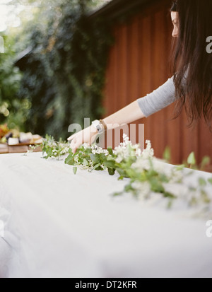 A woman with long hair by table laid outside with a white cloth and central foliage table decoration Place settings