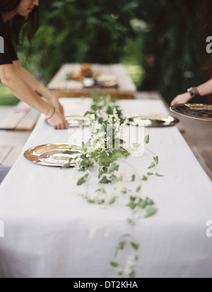 Two people leaning over a table laid outside with a white cloth and a central foliage table decoration Stock Photo