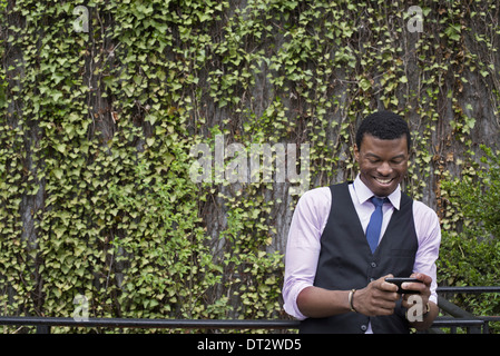 City park wall covered in climbing plants and ivy A young man in a waistcoat shirt and tie checking his phone Stock Photo