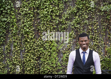 City park with a wall covered in climbing plants and ivy A young man in a waistcoat shirt and tie Looking at the camera Stock Photo