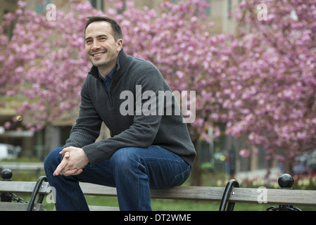 A man sitting on the back of a park bench under the cherry blossom trees in the park Stock Photo