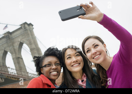 New York city the Brooklyn Bridge East River Three women in a row smiling as one takes a picture with a smart phone Stock Photo
