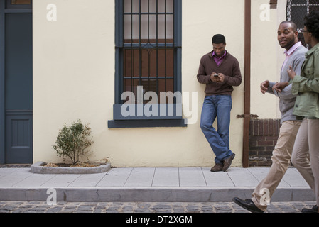 Outdoors in the city in spring A couple walking on the sidewalk and a man leaning against a wall checking his phone Stock Photo