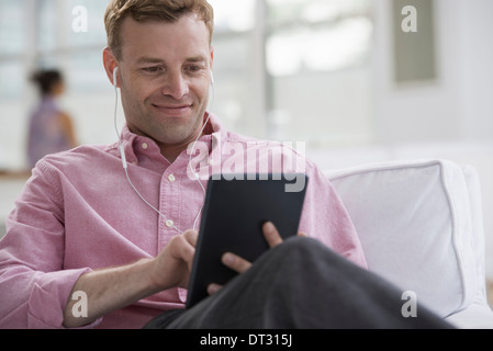 A man in a pink shirt sitting smiling using a digital tablet Wearing earphones Stock Photo