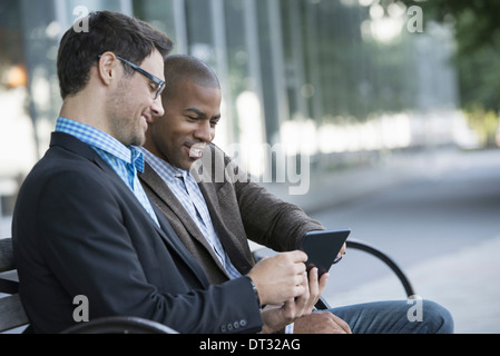 Two men seated on a park bench outdoors looking at a digital tablet Stock Photo