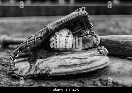 Baseball glory days with ball in glove and bat on base on field. Monochrome image with outfield wall in background. Stock Photo