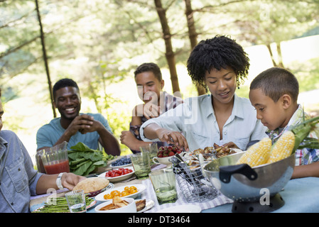 A family picnic in a shady woodland Adults and children around a table handing around plates and food Stock Photo