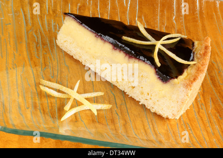 Slice of blueberry jam and custard cream tart served on a glass plate with lemon zest, over an orange background. Stock Photo