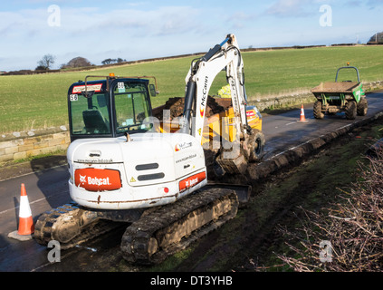 A Bob Cat backhoe excavator and Dumper Truck excavating a pipe trench along a rural road Stock Photo