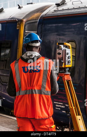 Surveyor standing next to his measuring equipment on a railway station platform with train in the background Stock Photo