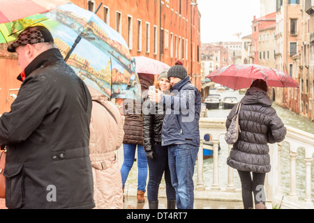 Venice, Italy. Young couple in winter and rain clothing taking pictures of themselfs at a rainy day on a bridge over a canal. Stock Photo