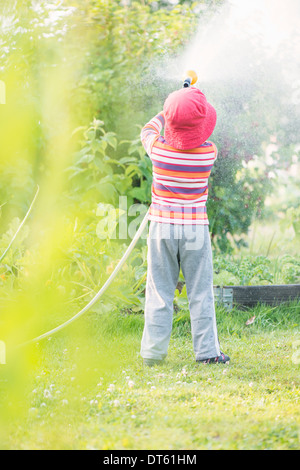 Lifestyle summer scene. Little girl watering garden plants and vegetables with sprinkler. Stock Photo