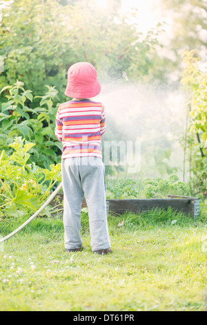 Lifestyle summer scene. Little girl watering garden plants and vegetables with sprinkler. Stock Photo
