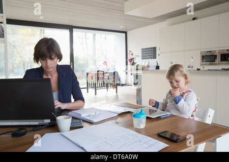 Mother with female toddler working on computer at kitchen table Stock Photo