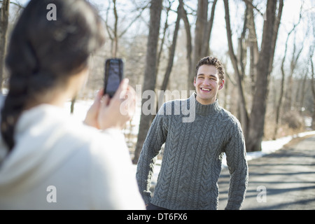 A couple outdoors on a snowy day. A woman holding a camera phone, taking photographs of a man. Stock Photo