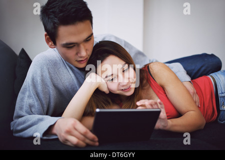 Lovely teenage couple lying together on sofa using tablet PC. Mixed race couple relaxing on couch surfing on the Internet. Stock Photo