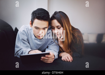 Teenage couple surfing internet on digital tablet while lying on sofa at home. Mixed race couple using tablet couple on couch. Stock Photo