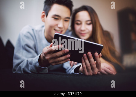 Digital tablet in young man's hand lying with his girlfriend on couch. Teenage couple looking at tablet PC. Mixed race couple. Stock Photo
