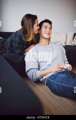 Handsome young man playing video game while his girlfriend whispering something in his ears. Teenage asian couple relaxing. Stock Photo