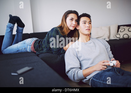 Beautiful teenage couple sitting together in living room at home. Young man playing video game with woman lying on sofa. Stock Photo