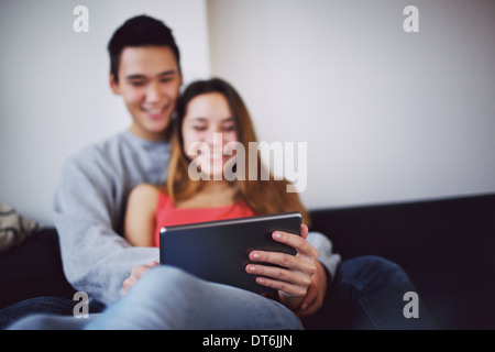 Happy teenage couple sitting comfortably on couch at home watching movie on digital tablet. Selective focus on tablet PC. Stock Photo