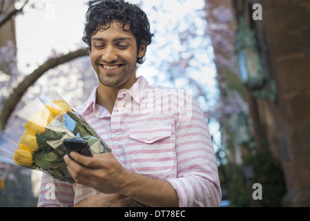 City life. A young man in the park in spring, using a mobile phone.  Holding a bunch of yellow roses. Stock Photo