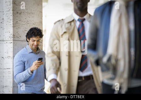 Three people on the sidewalk, a man using his mobile phone, a man in a raincoat, and a young man walking along the street. Stock Photo