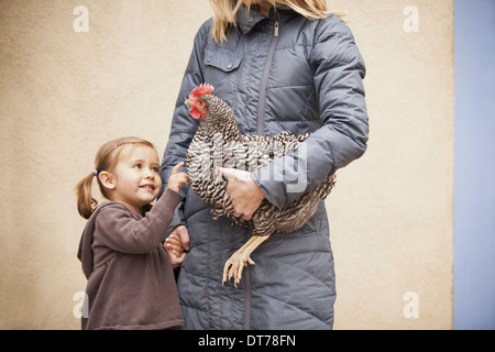 A woman in a grey coat holding chicken with a red coxcomb under one arm. A young girl beside her holding her other hand Stock Photo