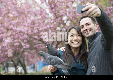 A couple, a man and woman, in the park, taking a selfy, self portrait with a mobile phone. Two pigeons perched on her wrist. Stock Photo