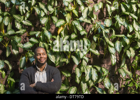 A man standing with folded arms, by a wall covered with foliage. Stock Photo