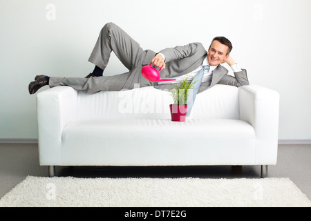 Photo of happy man watering plant while lying on sofa Stock Photo
