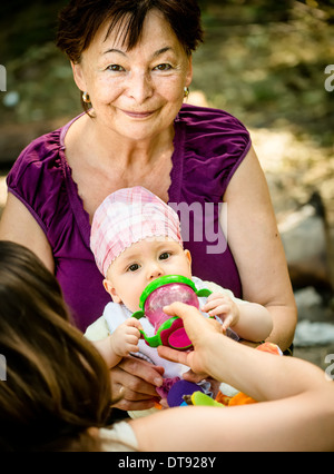 Mother gives a drink from bottle to her baby, grandmother is helping Stock Photo
