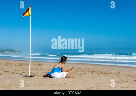 Woman sitting on the beach using her mobile phone, Sedgefield, Eastern Cape, South Africa Stock Photo