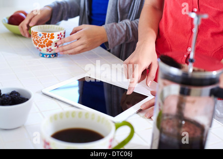 Two young women in kitchen using digital tablet whilst having coffee Stock Photo