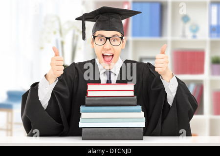 Excited college graduate giving thumbs up seated at table behind a stack of books, indoors Stock Photo
